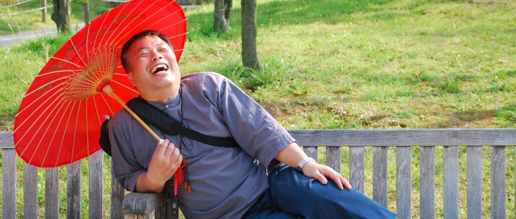 WHO-Empfehlungen: Ein lachender Mann sitzt unter einem Sonnenschirm auf einer Parkbank